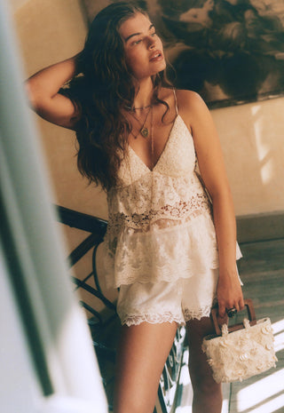 A woman wearing a white lace camisole top and matching lace shorts, holding a small white floral handbag in a sunlit indoor setting.