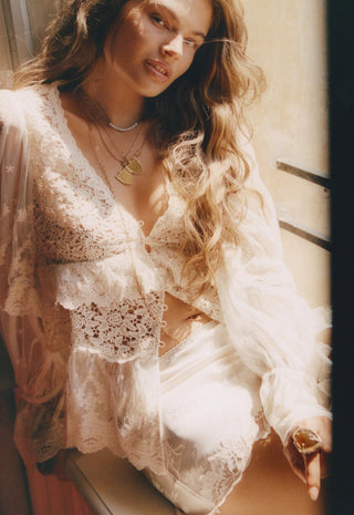 A woman wearing a white lace lingerie set with a sheer long-sleeve robe, styled with layered gold necklaces, in warm, sunlit indoor lighting.