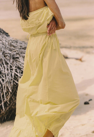 a-woman-wearing-a-flowing-pale-yellow-strapless-maxi-dress-standing-on-a-sandy-beach-next-to-a-thatched-structure