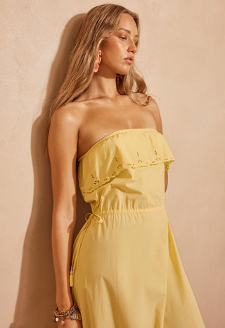 a-woman-wearing-a-strapless-pale-yellow-dress-with-eyelet-lace-detailing-on-the-bodice-standing-against-a-textured-beige-wall