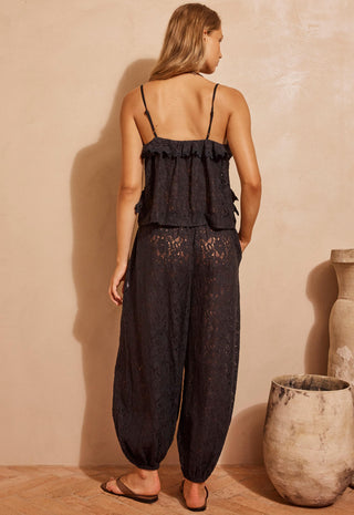 a-woman-stands-with-her-back-to-the-camera-wearing-a-black-lace-camisole-top-with-thin-straps-and-matching-black-lace-harem-pants-photographed-in-a-warm-beige-studio-setting-with-decorative-vases