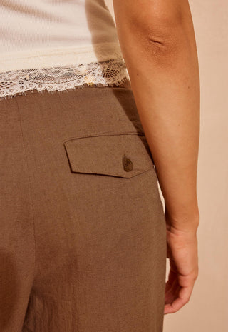 close-up-of-a-person-wearing-brown-wide-leg-trousers-with-a-flap-pocket-and-button-paired-with-a-cream-lace-trim-top-against-a-warm-beige-background