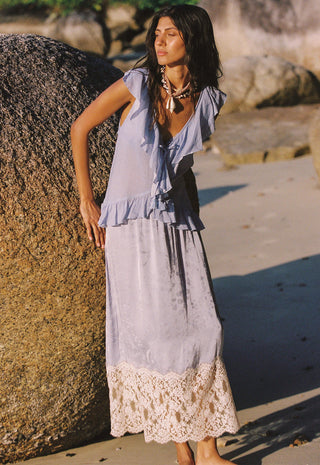 a-woman-stands-outdoors-on-a-sandy-beach-next-to-a-large-rock-wearing-a-light-lavender-sleeveless-maxi-dress-with-ruffled-shoulders-and-a-cream-lace-hem