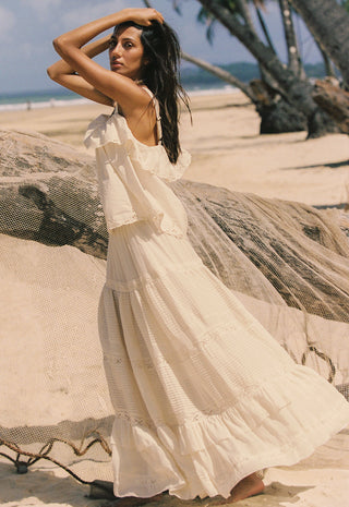 a-woman-wearing-a-flowing-cream-boho-style-maxi-dress-stands-on-a-sandy-beach-next-to-a-large-driftwood-log-with-palm-trees-and-the-ocean-in-the-background